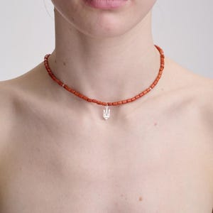 May include: A choker necklace featuring small, cylindrical, orange-red beads strung together with a central silver-toned pendant shaped like the Ukrainian Tryzub coat of arms. The necklace sits closely around the neck with a simple, uniform design.