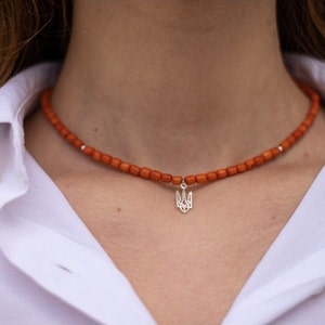 May include: A necklace featuring small, cylindrical orange beads strung together with a central silver-toned pendant shaped like a trident symbol. The beads are uniform in size and color, creating a consistent circular pattern around the neck.