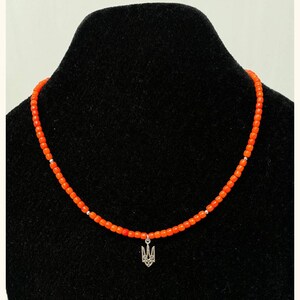 May include: A beaded necklace featuring small, vibrant orange cylindrical beads strung together with occasional silver-toned spacers. A silver-toned metal pendant shaped like the Ukrainian Tryzub emblem hangs from the center of the necklace.