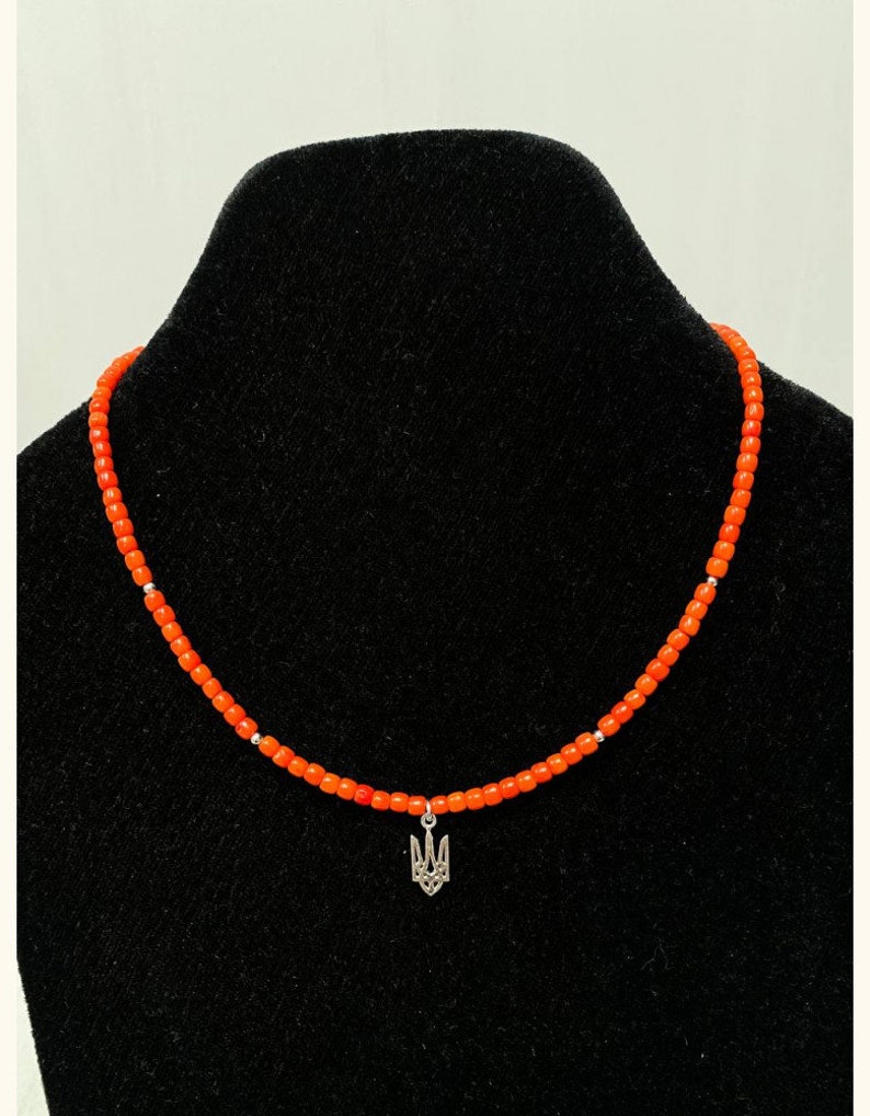 May include: A beaded necklace featuring small, vibrant orange cylindrical beads strung together with occasional silver-toned spacers. A silver-toned metal pendant shaped like the Ukrainian Tryzub emblem hangs from the center of the necklace.