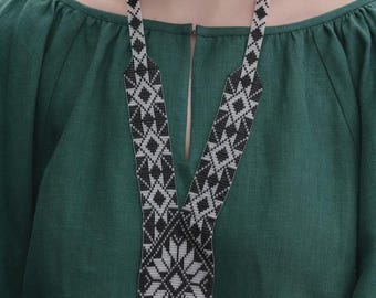 May include: A long beaded necklace featuring a geometric pattern in black and white. The design consists of repeating diamond and star-like motifs, ending in a pointed pendant section with a fringe of black beads at the bottom edge of the piece.