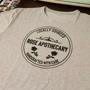 May include: A gray t-shirt with a black circular logo that says "Locally Sourced Rose Apothecary Handcrafted With Care". The logo features two stylized roses.