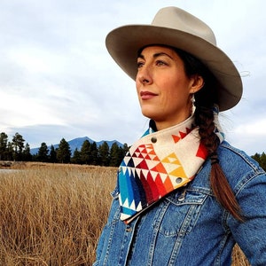 May include: A woman wearing a denim jacket with a colorful patterned scarf and a gray cowboy hat. She is standing in a field with tall grass and mountains in the background.