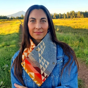 May include: A woman wearing a denim jacket and a geometric patterned neck warmer. The neck warmer features orange, red, and cream colours with a button closure. The background shows a dirt path, green field, and a mountain.