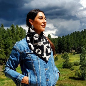 May include: A woman wearing a black and white geometric patterned scarf with a denim jacket. She is standing in a field with trees in the background.