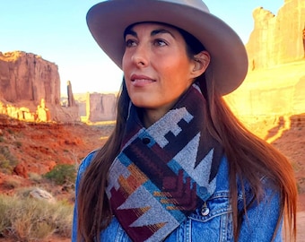 May include: A woman wearing a grey cowboy hat and a blue denim jacket with a colourful patterned neck warmer. The woman is standing in a desert landscape with red rock formations in the background.