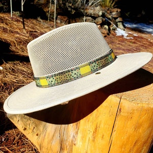 May include: A beige mesh hat with a wide brim and a patterned band. The band is made of a fabric with a yellow, green, and brown pattern. The hat is sitting on a wooden stump.