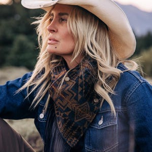 Puede incluir: Un sombrero de vaquero de paja color crema usado con una chaqueta de mezclilla azul oscuro y un pañuelo de tonos oscuros con un patrón geométrico en marrón y negro, con botones de presión metálicos y flecos negros largos.