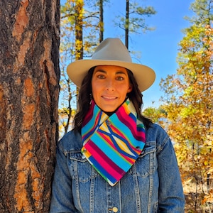 Puede incluir: Un sombrero de ala ancha gris claro y un calentador de cuello colorido con rayas vibrantes en rosa, azul, amarillo y morado. El calentador de cuello se asegura con dos botones de presión metálicos y se lleva sobre una chaqueta vaquera azul.