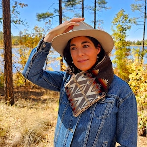 May include: A denim jacket with a beige hat and a patterned neck warmer. The neck warmer features a geometric design in shades of brown, beige, red, and orange. The background includes trees and a lake, suggesting an outdoor setting.