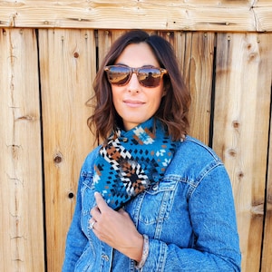 Model is standing in front of wooden door, wearing western cowl scarf.  Bandana scarf is made from wool & cotton, features blue, white, black & brown geometric designs.