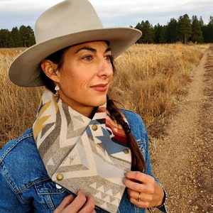 May include: A woman wearing a denim jacket and a gray and white patterned scarf with gold buttons. She is wearing a gray cowboy hat and has long brown hair. The background is a field of dry grass and a dirt road.
