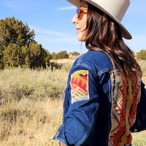 May include: A woman wearing a blue denim jacket with colorful patterned fabric accents on the back and sleeve. She is wearing a white hat and sunglasses and is standing in a field.