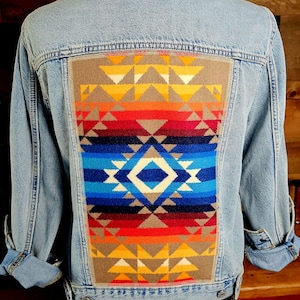 May include: Light blue denim jacket with a vibrant, geometric patterned back panel. The panel features a colorful design with shades of yellow, orange, red, blue, and brown. The jacket has button closures and long sleeves.