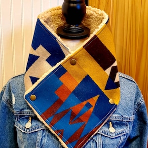 May include: A blue, yellow, orange, and brown patterned fleece neck warmer with a white sherpa lining. The neck warmer is folded in half and is being displayed on a mannequin.