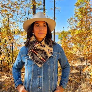 May include: A beige cowboy hat and a patterned neck gaiter with a button closure are worn over a denim jacket. The neck gaiter features a geometric design in brown, beige, red, and turquoise. The background shows a forest with fall foliage.