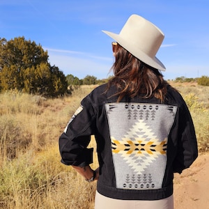 May include: Black denim jacket with a geometric design on the back in gray, yellow, and white. The jacket has long sleeves and a classic collar. A light-colored hat is worn.