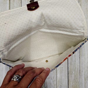 May include: Open clutch purse with a floral pattern in red, blue, and cream. The interior is a cream color with a small cross pattern. The purse has a brown leather strap and a brass clasp.