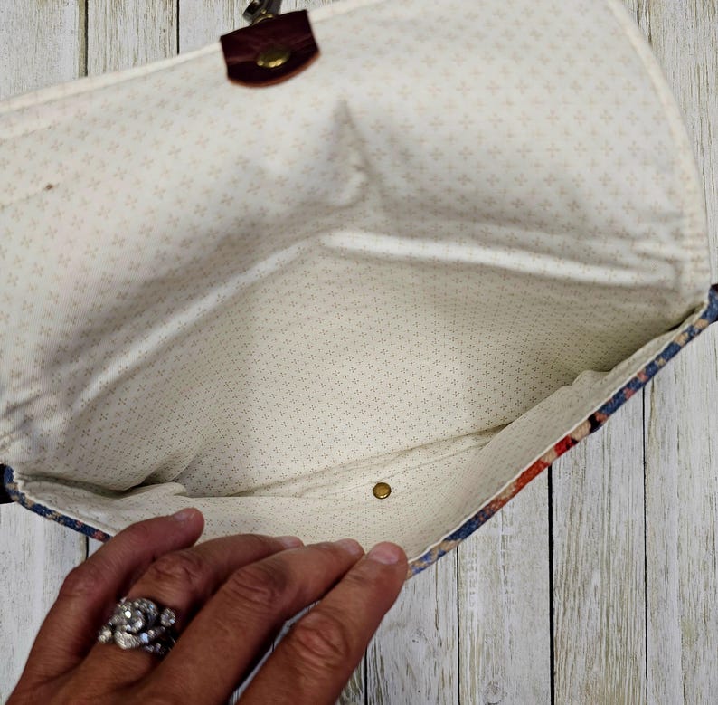 May include: Open clutch purse with a floral pattern in red, blue, and cream. The interior is a cream color with a small cross pattern. The purse has a brown leather strap and a brass clasp.