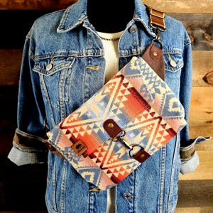 May include: A crossbody bag featuring a geometric pattern in blue, orange, and cream. It has brown leather details, a metal clasp, and a brown leather strap. The bag is shown against a denim jacket.