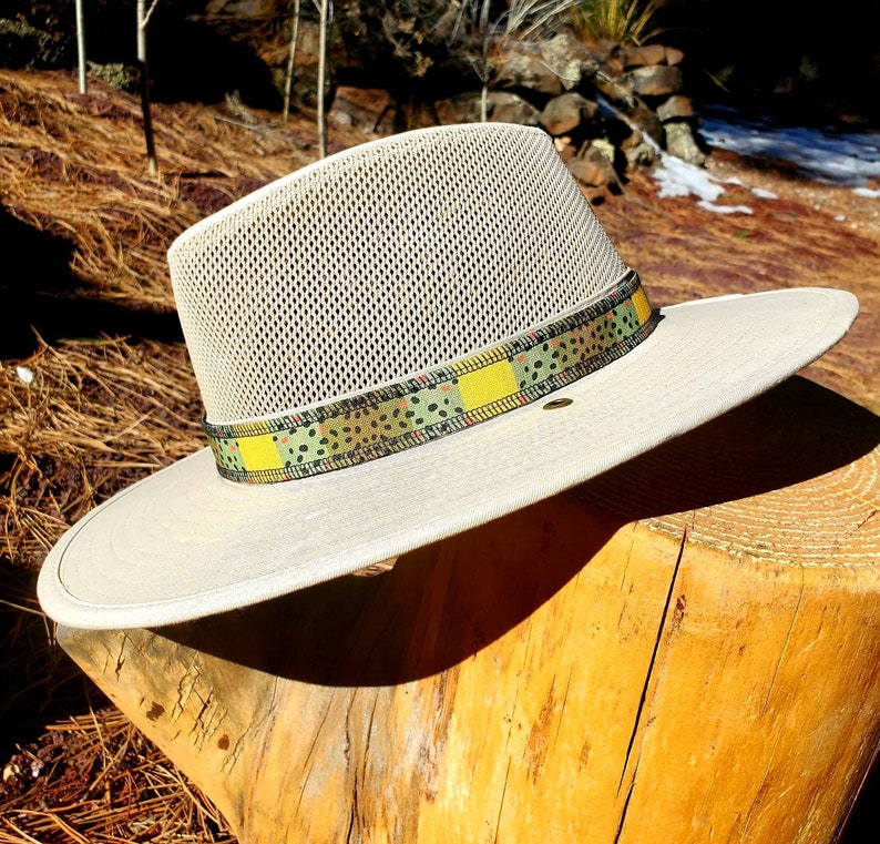 Light beige river hat leaning on wooden stump, featuring brown trout patterned hat band.  Hat band has yellow, green, brown coloring with black speckles mimicking the coloring of brown trout.