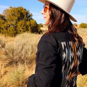 May include: A black denim jacket featuring a large, rectangular back panel with a woven geometric pattern in shades of blue, white, black, and yellow. The jacket is worn with a light-colored fedora hat and orange-tinted sunglasses.