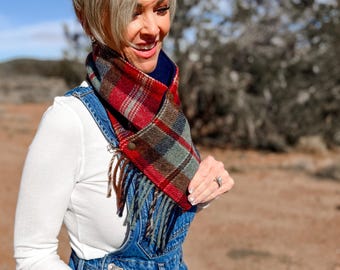 May include: A plaid neck warmer featuring a red, green, and brown tartan pattern with a dark blue interior lining. The accessory is secured with a metallic snap button and finished with a fringe trim along the bottom edge for a textured look.