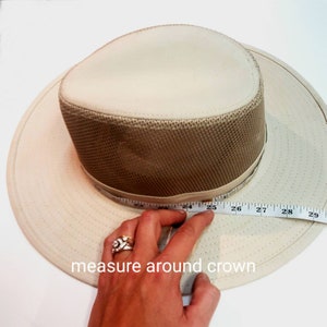 May include: A beige hat with a brown mesh crown and a white measuring tape around the crown. The tape reads "24, 25, 26, 27, 28, 29". The text "measure around crown" is written below the hat.