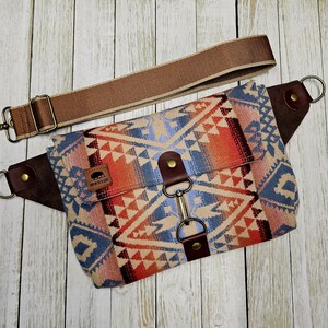 May include: A patterned crossbody bag with a brown adjustable strap and leather accents. The bag features a geometric design in shades of blue, orange, and beige. The bag has a metal clasp and a small logo patch.