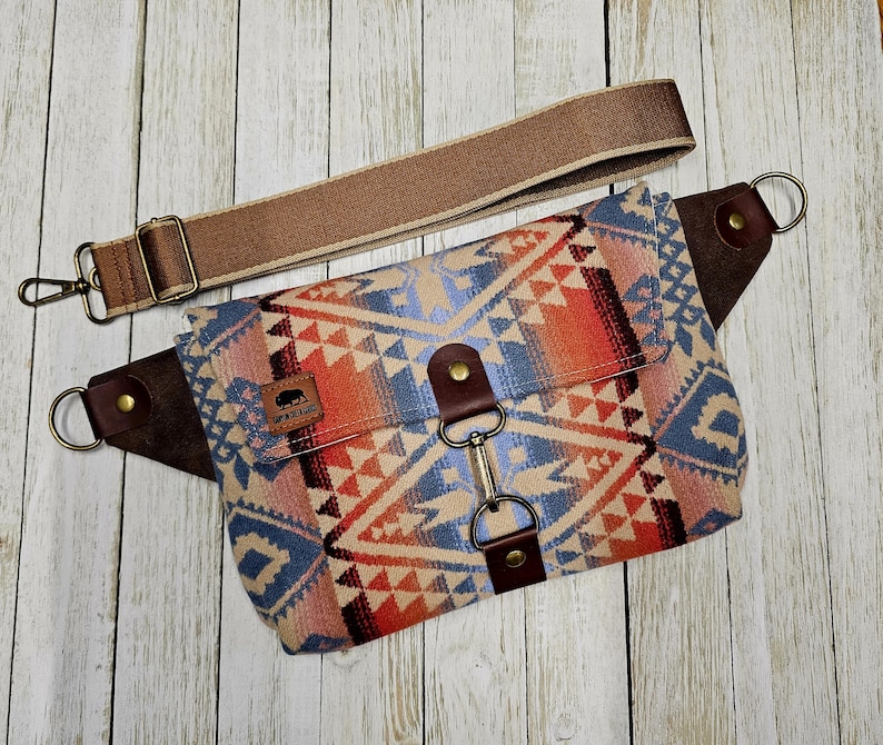 May include: A patterned crossbody bag with a brown adjustable strap and leather accents. The bag features a geometric design in shades of blue, orange, and beige. The bag has a metal clasp and a small logo patch.