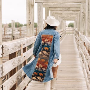 May include: A long blue denim jacket featuring a wide back panel with a colorful woven pattern of bison and geometric shapes. The jacket has long sleeves, a frayed hem, and is worn with a white cowboy hat and tall light-colored boots.