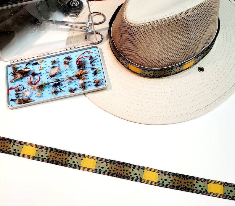 May include: A brown and tan patterned ribbon with yellow accents. The ribbon is laid out on a white surface. A fishing hat with a brown and tan patterned band is in the background.