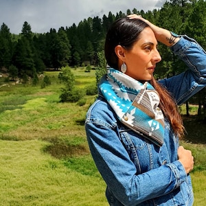 May include: A woman wearing a blue denim jacket with a turquoise, white, and brown patterned scarf. The scarf is folded over and tied at the neck. The woman is standing in a field with a forest in the background.