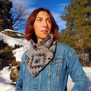 May include: A woman wearing a denim jacket with a gray and brown patterned scarf. The scarf has a button closure and is folded over to create a cowl neck style. The woman is standing in a snowy landscape with trees and rocks in the background.