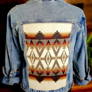 May include: Light blue denim jacket with a colorful geometric pattern on the back. The pattern features diamonds and triangles in shades of brown, red, and cream, creating a Southwestern or Native American-inspired design. The jacket has long sleeves and a classic collar.