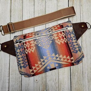 May include: A colorful fanny pack with a geometric pattern in shades of blue, orange, and beige. It features a white zipper, brown leather accents, and a detachable brown strap with metal hardware. The bag is designed for carrying essentials.