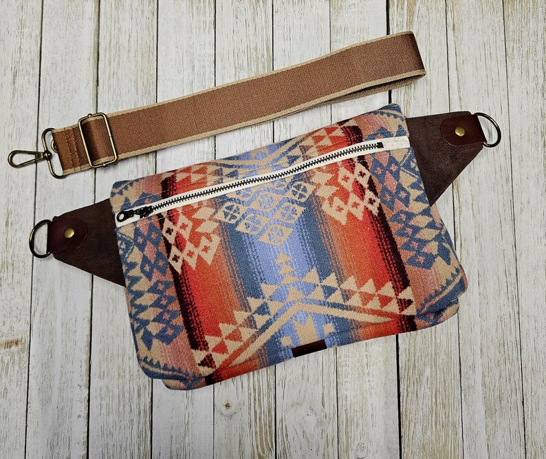 May include: A colorful fanny pack with a geometric pattern in shades of blue, orange, and beige. It features a white zipper, brown leather accents, and a detachable brown strap with metal hardware. The bag is designed for carrying essentials.