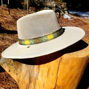 May include: A beige mesh hat with a wide brim and a patterned band in shades of green, yellow, and black. The hat is sitting on a wooden stump.