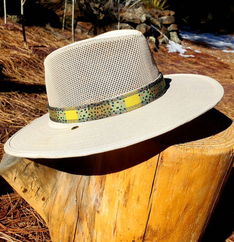 May include: A beige mesh hat with a wide brim and a patterned band in shades of green, yellow, and black. The hat is sitting on a wooden stump.