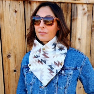 May include: A white and brown geometric patterned neck warmer with brown button accents. The neck warmer is worn by a person wearing a denim jacket and sunglasses.