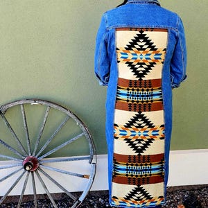 May include: A long denim jacket with a colorful geometric pattern on the back. The pattern includes black, brown, blue, and orange shapes. A white hat is worn.
