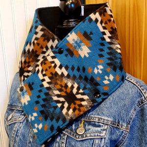 Puede incluir: Un calentador de cuello de forro polar azul, marr&oacute;n, negro y blanco con botones negros. El calentador de cuello est&aacute; doblado y se muestra en una chaqueta vaquera.