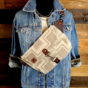 May include: A beige and white patterned crossbody bag with brown leather details and a metal clasp. The bag is worn over a denim jacket and a white top. The bag features a geometric design and a small leather patch.