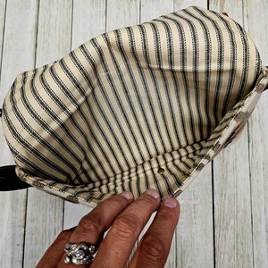 Puede incluir: Vista interior de una bolsa con tela a rayas beige y negras. La bolsa tiene una correa de cuero marr&oacute;n y un exterior estampado. La tela tiene rayas negras verticales sobre un fondo beige. Se ve una mano.