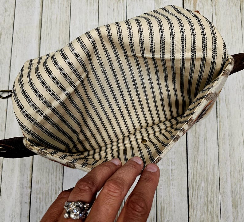 Puede incluir: Vista interior de una bolsa con tela a rayas beige y negras. La bolsa tiene una correa de cuero marr&oacute;n y un exterior estampado. La tela tiene rayas negras verticales sobre un fondo beige. Se ve una mano.
