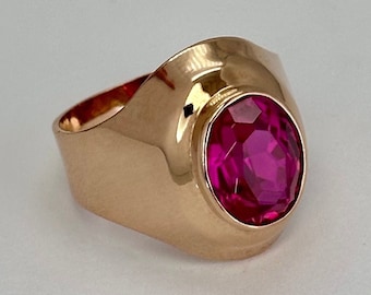 May include: A gold ring with a large, round, pink gemstone set in a raised bezel. The ring has a wide band with a rounded, curved top.