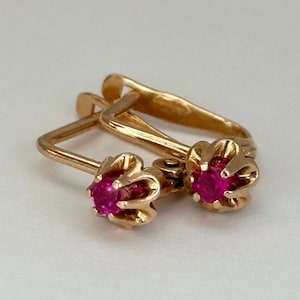 Op de afbeelding: Een paar gouden oorbellen met roze edelstenen. De oorbellen hebben een eenvoudig ontwerp met een kleine, ronde edelsteen die in een gouden zetting is gezet.