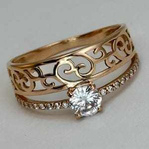 May include: A gold ring with a delicate filigree design and a single, clear, round gemstone in the center. The ring is set with smaller, clear gemstones around the band.