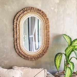 Könnte beinhalten: Ovaler Spiegel mit einem natürlichen Webrahmen. Der Rahmen hat ein Fransen-Design und kleine Holzperlen. Der Spiegel ist an einer hellgrauen Wand angebracht, mit einer grünen Pflanze in einem geflochtenen Korb rechts.