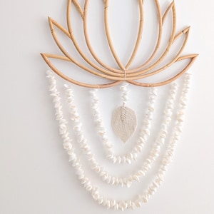 May include: A natural-toned, lotus-shaped wall hanging crafted from woven rattan. Below the lotus, strands of white seashells cascade downwards, with a central, textured leaf-shaped accent. This decorative piece adds a touch of bohemian style.